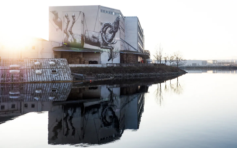 Großes Wandbild auf einem Industriegebäude, das sich im Wasser des Linzer Hafens spiegelt